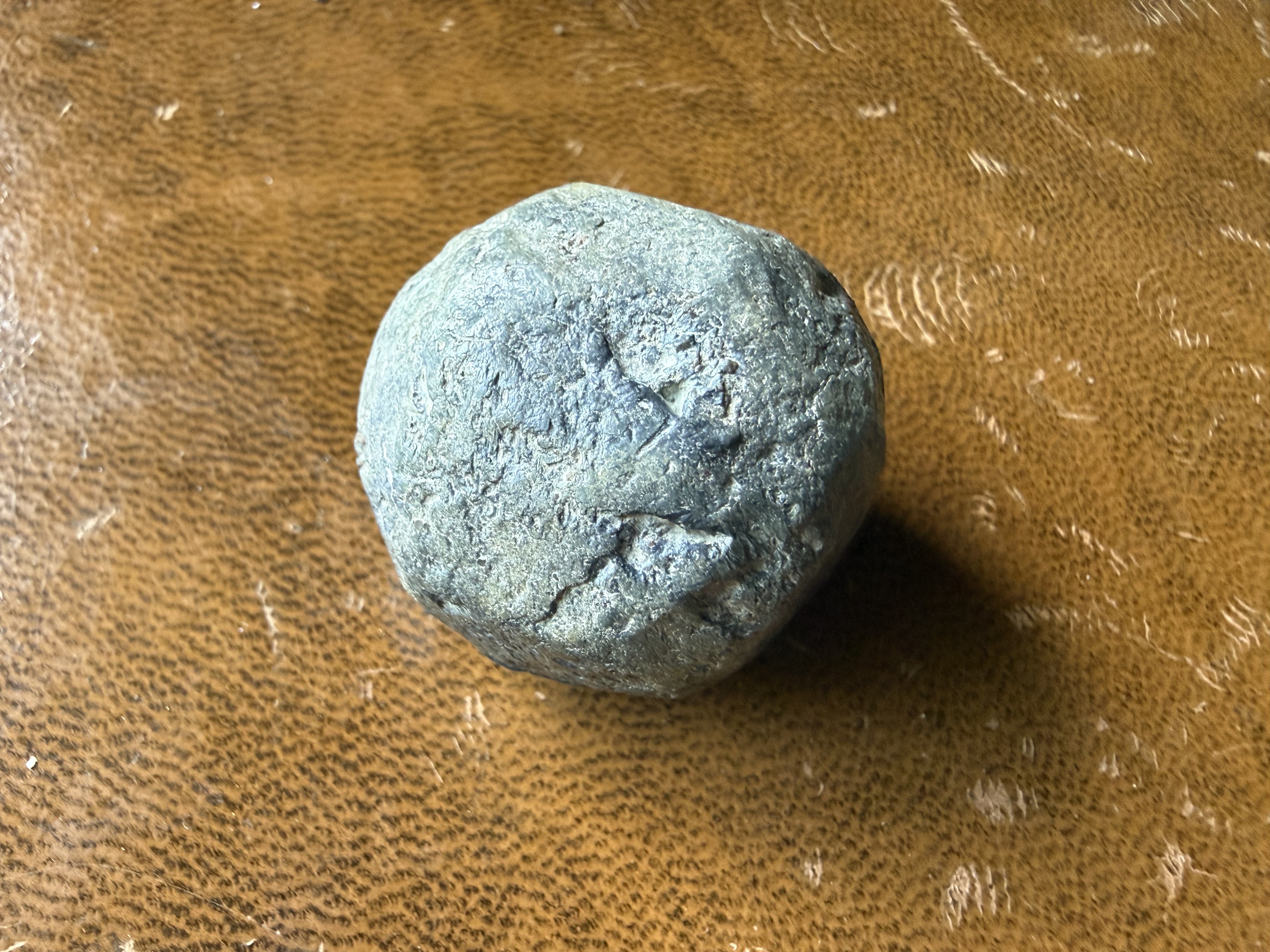 Heavily corroded spherical lead musket ball with green patination and surface cracking, displayed on brown leather, found by metal detecting